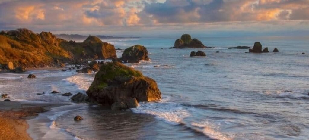 Rugged coastline of Luffenholtz Beach in Northern California, at sunset with large sea stacks, rocky coves, and glowing clouds over the Pacific Ocean.
