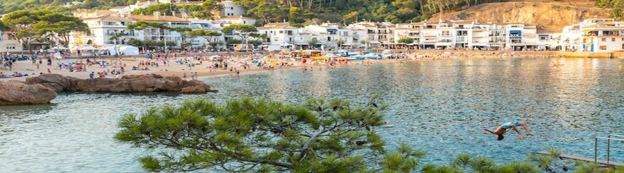 People swimming and relaxing along a lively cove beach in Costa Brava, Spain, surrounded by white villas and rocky cliffs.