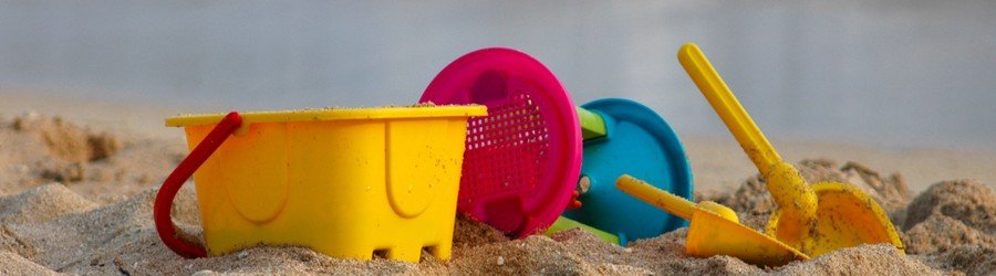 Colorful beach toys including a yellow bucket, pink sifter, and yellow shovel in the sand.