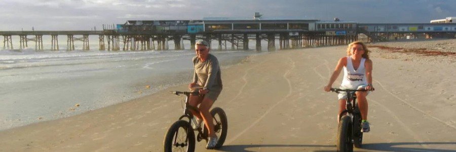 Visitors riding bicycles along the sandy shoreline near Cocoa Beach Pier in Florida.