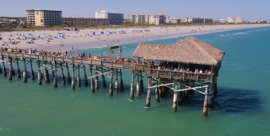 Aerial view of the historic Westgate Cocoa Beach Pier in Florida, featuring the thatched-roof Riki Tiki Tavern over turquoise ocean water with a crowded sandy beach and hotels in the background.