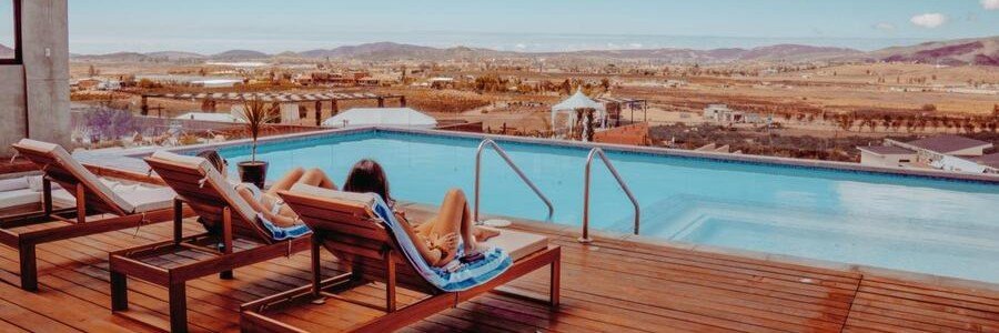 Three travelers relaxing on wooden lounge chairs by a rooftop infinity pool, overlooking the vast valley and mountains of Valle de Guadalupe.