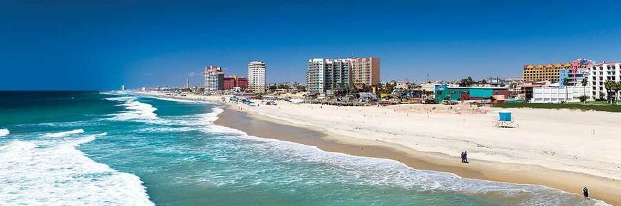 Aerial view of Playa Rosarito in Rosarito Mexico with sandy beaches, rolling ocean waves, coastal mountains, and beachfront homes along the Pacific coastline.