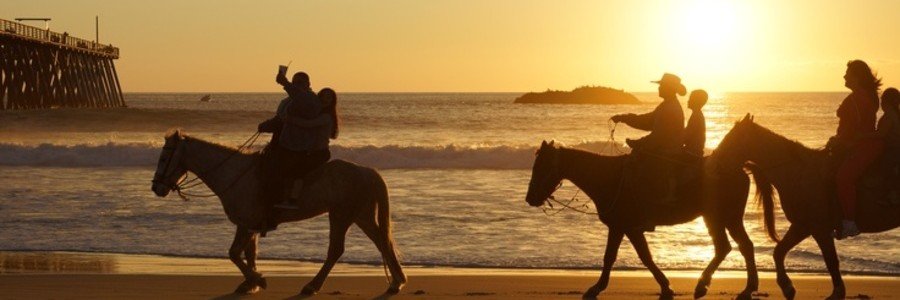 Silhouettes of horseback riders along the beach at sunset in Rosarito Mexico with golden ocean waves, sandy shoreline, and a nearby pier.