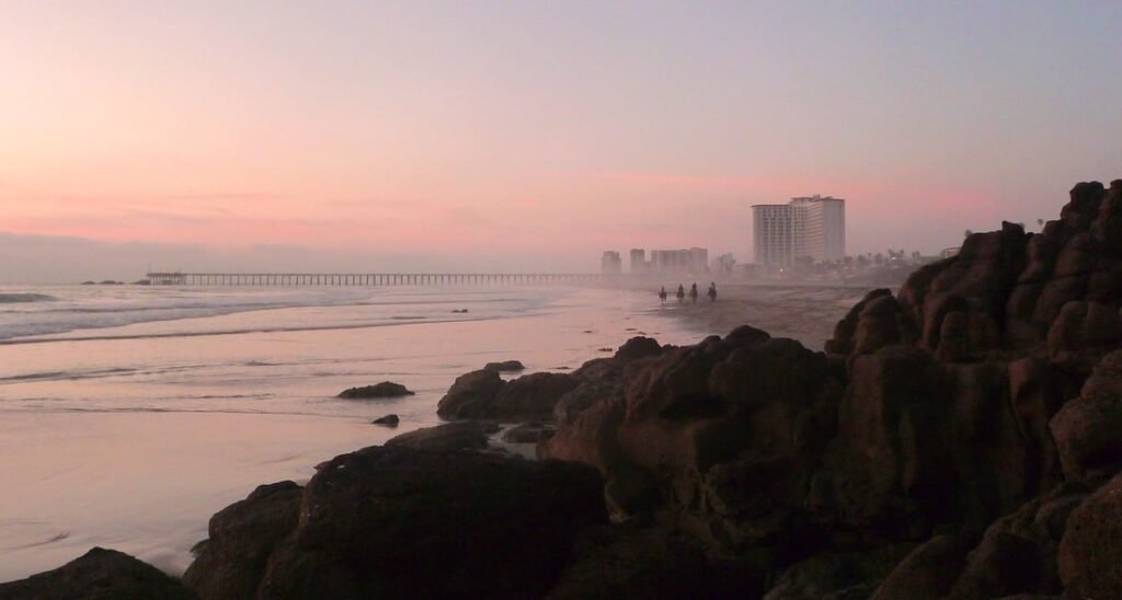 Sunset along the rocky coastline in Rosarito Mexico with soft pink skies, ocean waves, beach walkers, distant resorts, and a long pier fading into the coastal mist.