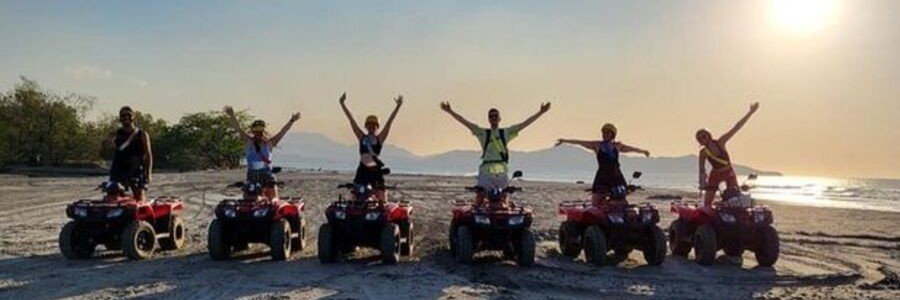 A group of travelers on red ATVs celebrating on the wide sandy beach of San Felipe at sunset with desert mountains in the background.