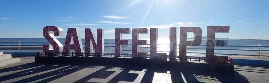 Large purple letters spelling 'SAN FELIPE' along a seaside promenade under bright sun and calm blue water in the background.