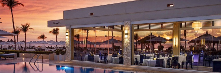 Bucuti Tara Beach Resort Elements Restaurant | Beach Travel Destinations Sunset at a resort poolside restaurant with outdoor tables, palm trees, and a glass-walled dining area.
