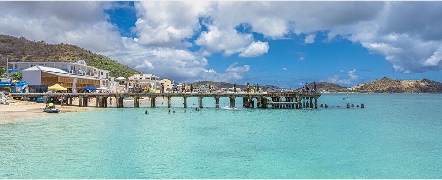 Grand Case Beach St. Martin with wooden pier, turquoise Caribbean water, beachfront restaurants, and hills under partly cloudy sky