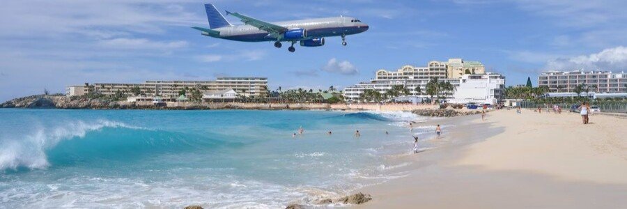 Airplane flying low over Maho Beach in St. Martin with turquoise water and white sand below.