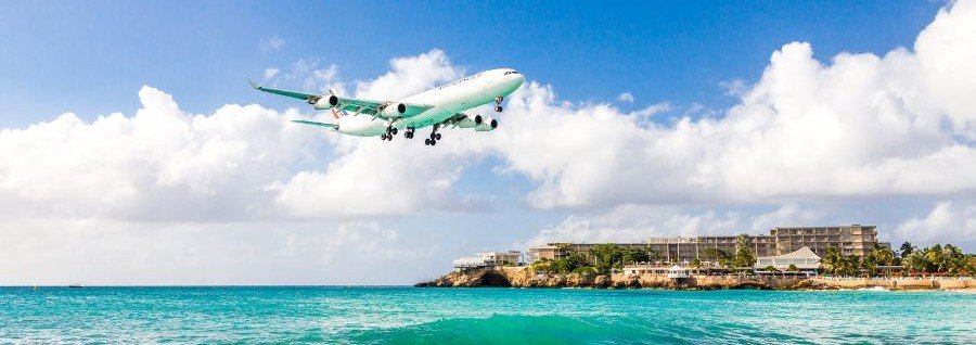 Airplane landing low over Maho Beach in St Martin with turquoise Caribbean water and coastal resort view.