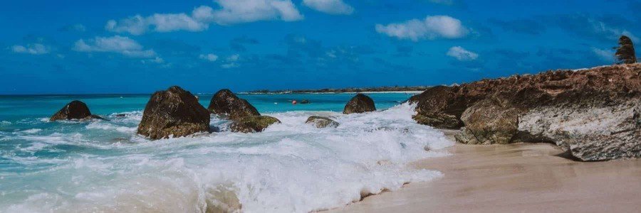 Rocky Coastline Ocean Waves | Beach Travel Destinations Ocean waves crashing onto a sandy beach shore with large dark volcanic rocks and a bright blue sky