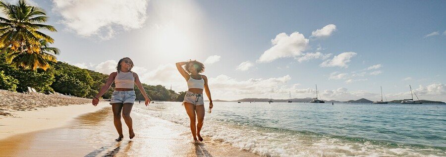 Two women walking along a white sand beach in St. Thomas with turquoise water, palm trees, and sailboats in the background under a sunny sky.