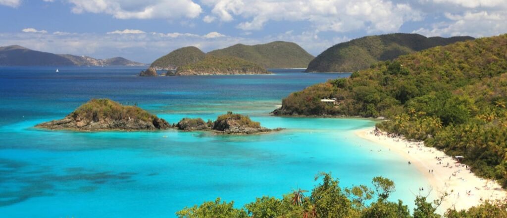 Aerial view of a turquoise bay in St. Thomas with a white sand beach, small islands, and lush green hills under a bright blue sky.