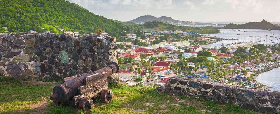 Historic cannon overlooking Philipsburg St. Martin harbor with colorful town buildings, green hills, and Caribbean coastline