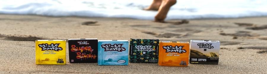 Sticky Bumps surf wax bars displayed on the beach sand with ocean waves in the background.
