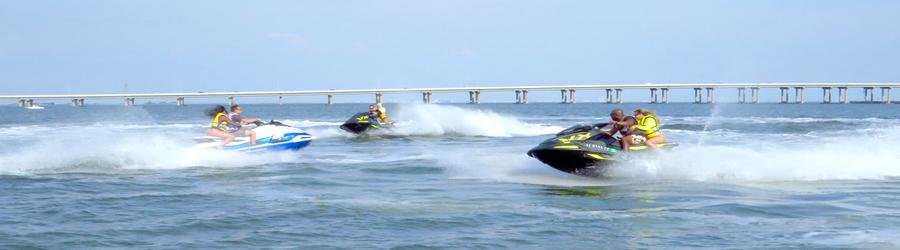 Group of people racing jet skis together on open water near a bridge.