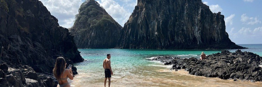 Exploring Baía dos Porcos Beach | Beach Travel Destinations Travelers exploring the sandy shoreline of Baía dos Porcos with the massive Morro Dois Irmãos rocks looming in the background.