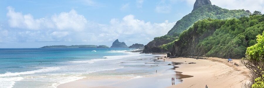 Panoramic view of Praia da Cacimba do Padre beach in Fernando de Noronha, Brazil, with the dramatic volcanic peak of Morro do Pico rising over the lush green cliffs.