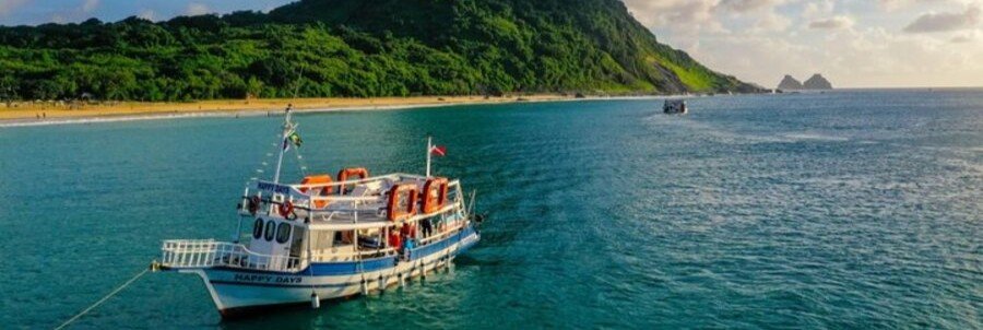 Traditional Tour Boat in Fernando de Noronha | Beach Travel Destinations White and blue tour boat anchored in calm turquoise waters alongside the lush, forested coastline of Fernando de Noronha.