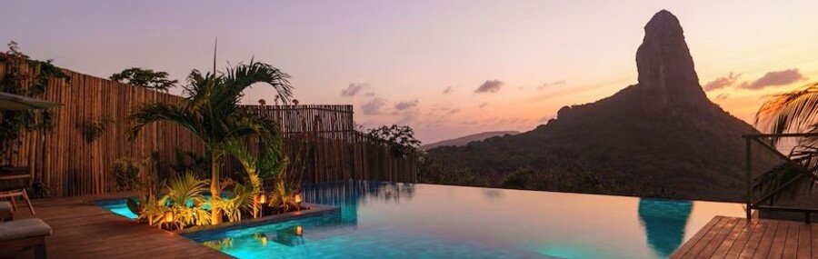 Infinity Pool Sunset at Pousada Ecológica Akanã | Beach Travel Destinations Infinity swimming pool and wooden sun deck at Pousada Ecológica Akanã during sunset, overlooking the dramatic volcanic peak of Morro do Pico.