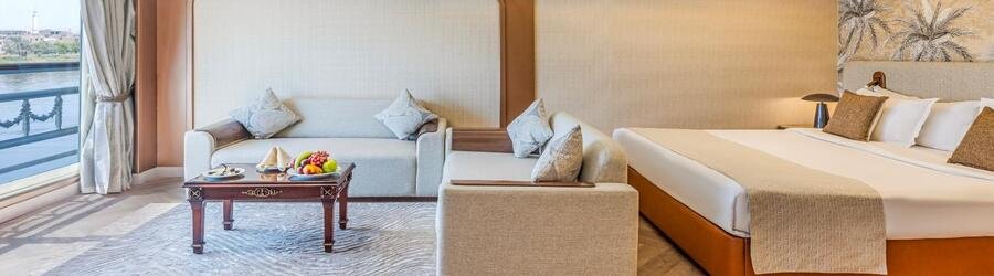 Luxury cruise cabin with king bed, sitting area, and panoramic ocean-view windows.