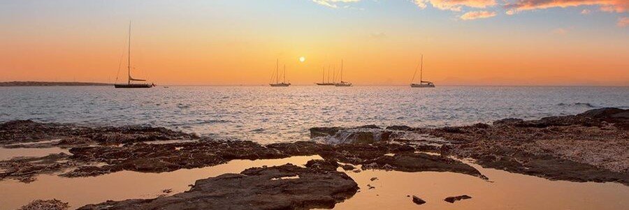 Sailboats anchored in the sea during a vibrant orange sunset viewed from the rocky shore of Cala Saona in Formentera.