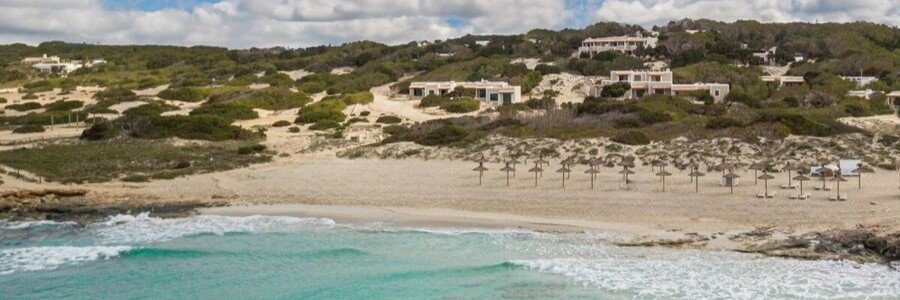 An aerial panoramic view of the sand dunes, straw beach umbrellas, and clear turquoise waters at Dunas de Formentera hotel along Migjorn beach.