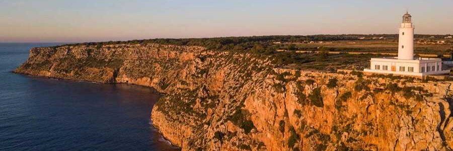 The historic white La Mola Lighthouse standing on the edge of high rocky sea cliffs in Formentera at sunset.