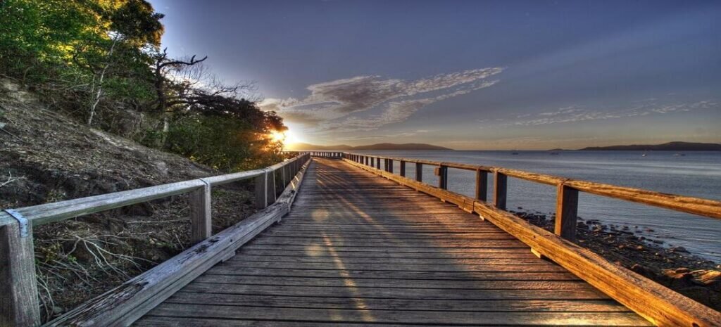 Wooden coastal boardwalk at sunrise overlooking calm water and hills in Australia’s Whitsunday Islands.