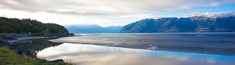 Wide view of Turnagain Arm near Anchorage with calm tidal flats, mountains, and soft morning light.