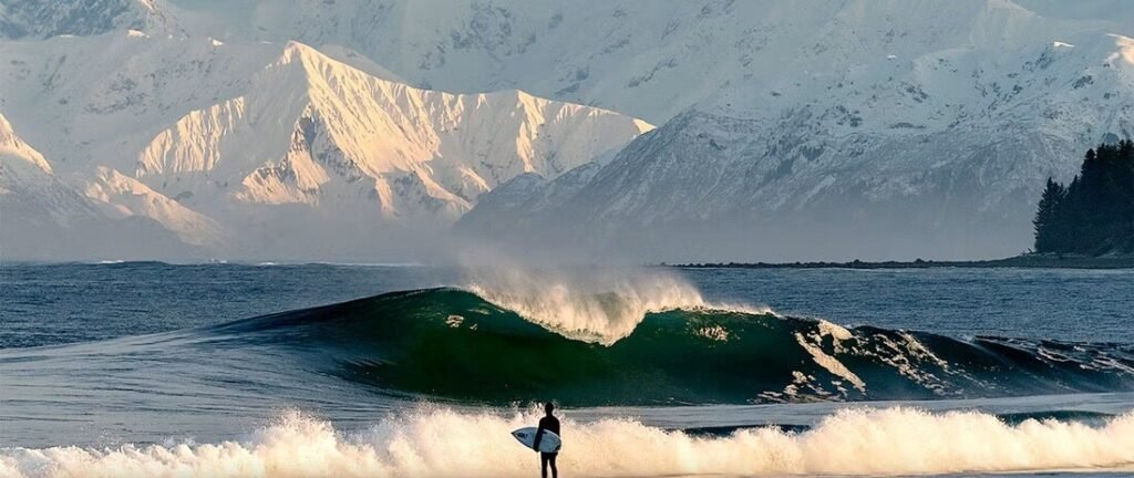 Surfer watching powerful ocean wave with snow-capped mountains near Yakutat Alaska coastline.