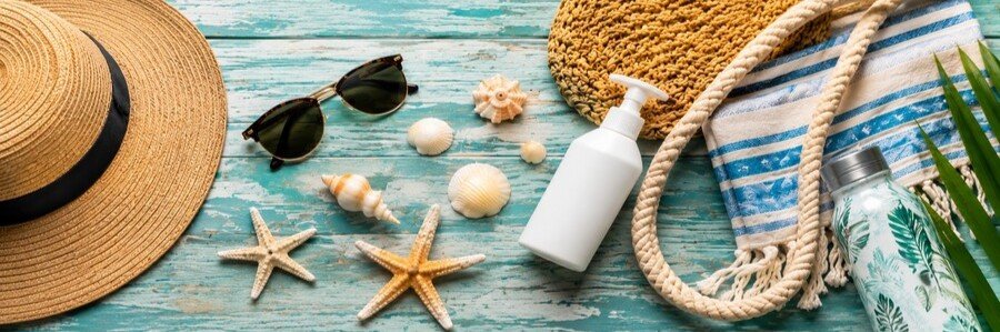 Beach vacation essentials including a sun hat, sunglasses, towel, sunscreen, shells, and reusable water bottle arranged on a wooden background.