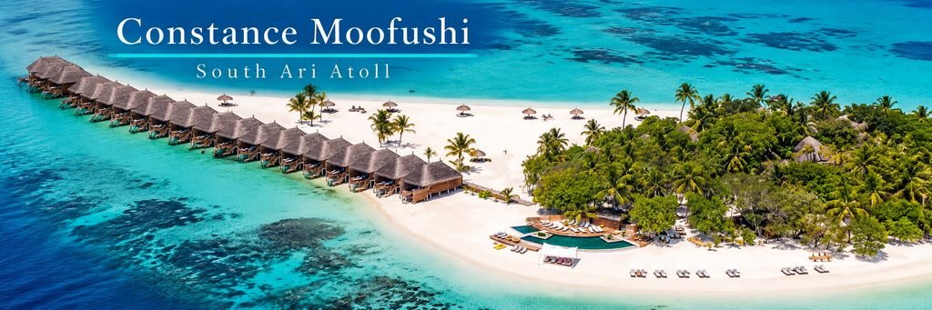 Aerial view of thatched-roof overwater villas beside a white-sand beach, surrounded by turquoise lagoons, coral reefs, and lush tropical palm trees.
