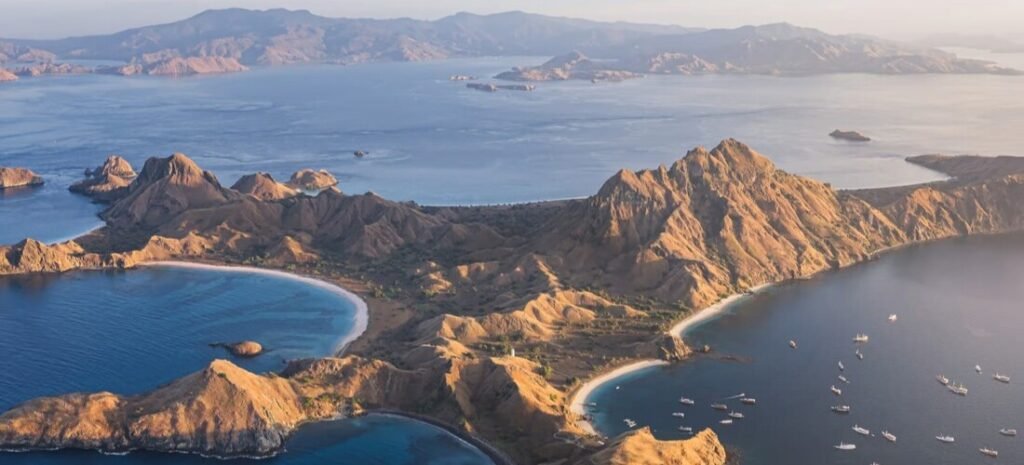Aerial View of Komodo Island | Beach Travel Destinations Aerial view of Komodo Island showing rugged hills, turquoise bays, white sand beaches, and boats anchored in Indonesia.