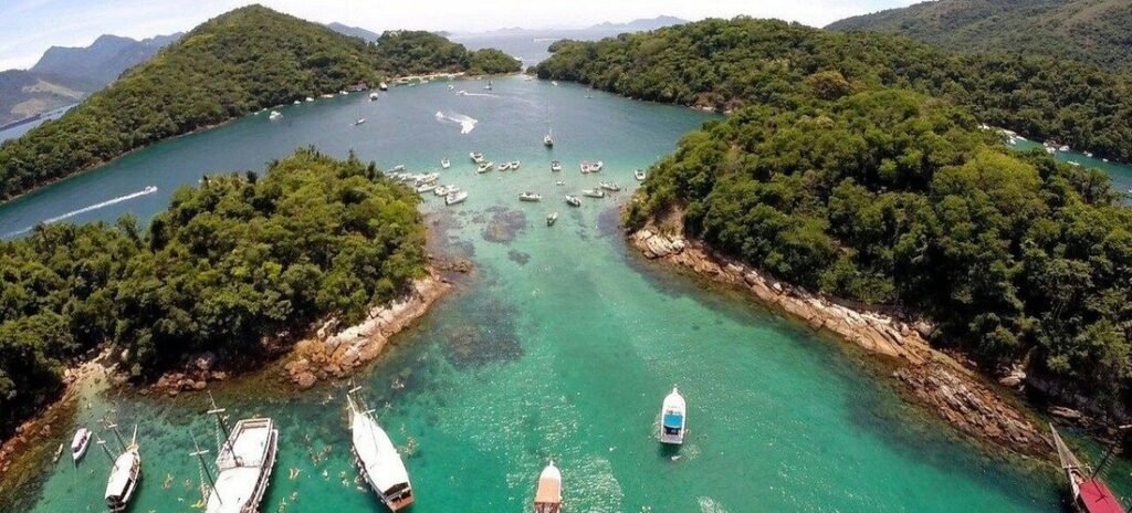 Aerial panorama of Lagoa Azul in Ilha Grande, Brazil, showing speedboats and schooners anchored in a narrow turquoise channel between lush, forested islands.