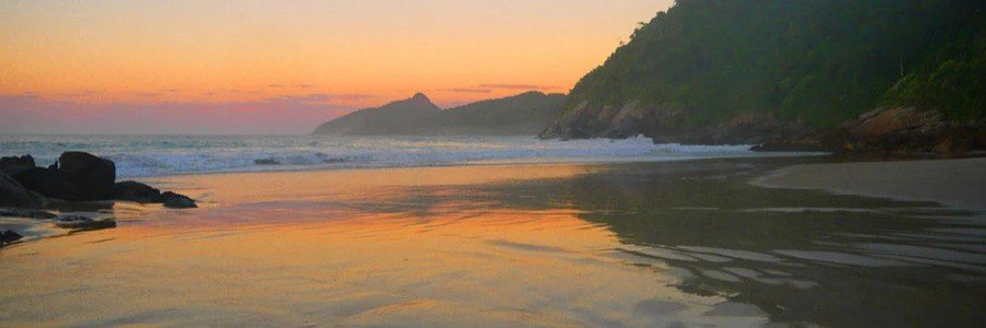Sunset over the wide sandy shore of Lopes Mendes Beach in Ilha Grande Brazil with orange sky reflections on the wet sand and forest mountains in the background