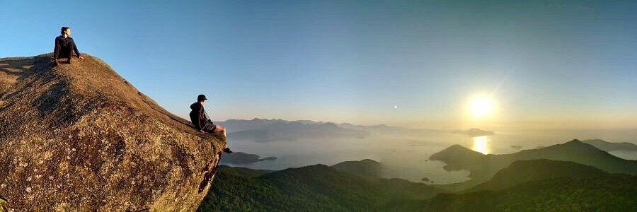 Two hikers sitting on a massive steep granite boulder at the summit of Pico do Papagaio looking out at the sun rising over the bays of Ilha Grande Brazil