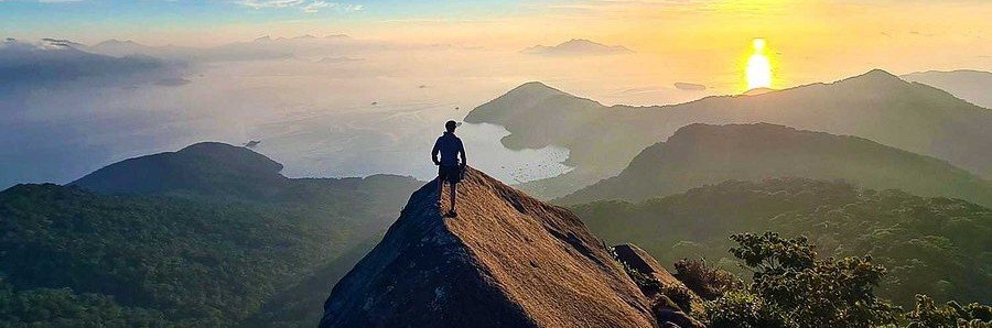Hiker standing on the steep granite summit ridge of Pico do Papagaio at sunrise overlooking the coast and mountains of Ilha Grande Brazil