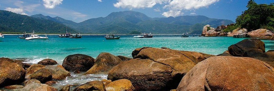 Large smooth boulders on the shore of Praia do Aventureiro in Ilha Grande Brazil with turquoise water and boats moored in the bay