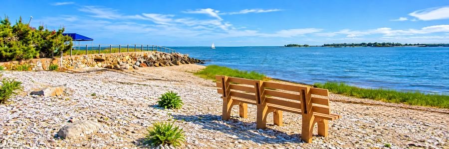 Canal Beach, one of the quieter Westport Beaches in Connecticut, featuring a pebbled shoreline, wooden bench, coastal greenery, and blue Long Island Sound waters.