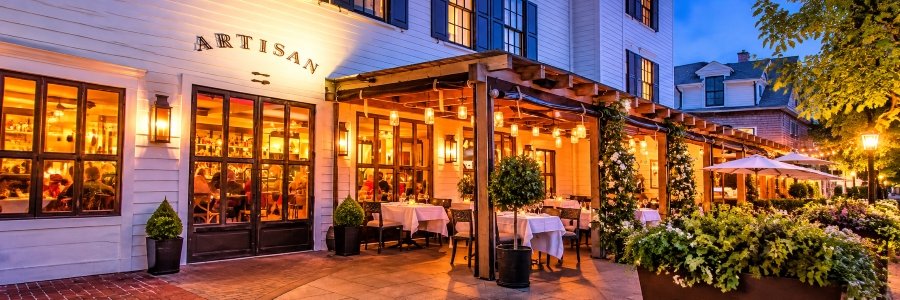 Elegant boutique hotel restaurant at dusk featuring a warmly illuminated outdoor dining terrace, white tablecloths, hanging lanterns, and lush greenery.