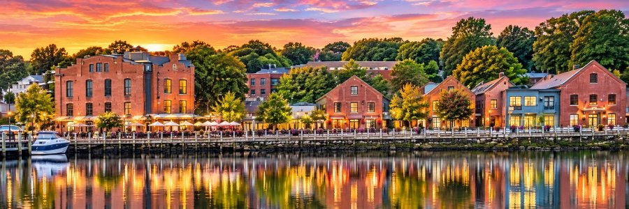 Beautiful sunset view of downtown Westport along the Saugatuck River, featuring historic brick buildings, waterfront dining, a docked boat, and colorful reflections on the water.