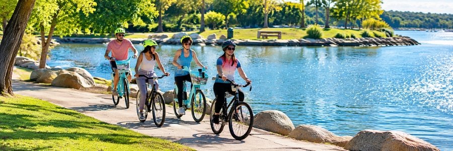 Four cyclists wearing helmets enjoy a scenic waterfront bike path surrounded by leafy trees, green lawns, large rocks, and sparkling blue water.