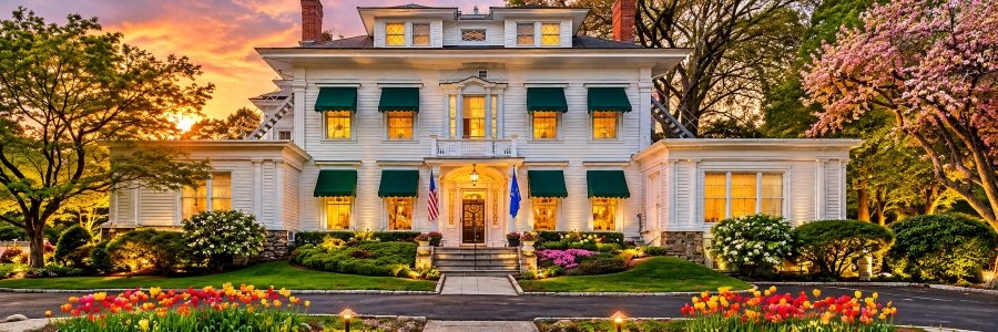 Elegant historic white mansion at sunset featuring green window awnings, warmly illuminated windows, colorful tulip gardens, and a grand entrance.