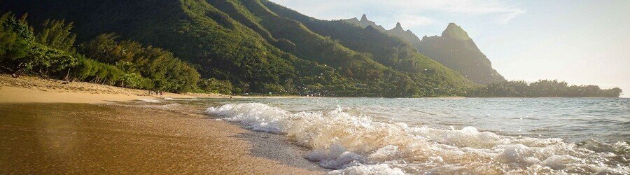 Keʻe Beach in Kauai with gentle waves, green mountains, and lush tropical scenery along the shoreline.