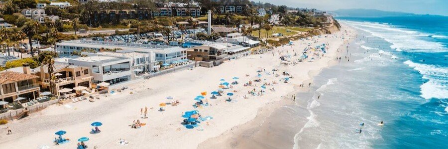 Aerial view of Del Mar Beach Hotel beside a wide sandy beach and rolling Pacific Ocean waves in Del Mar, California.