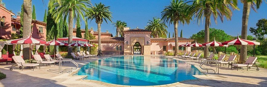 Large swimming pool at Fairmont Grand Del Mar surrounded by palm trees, lounge chairs, striped umbrellas, and Mediterranean-style buildings.