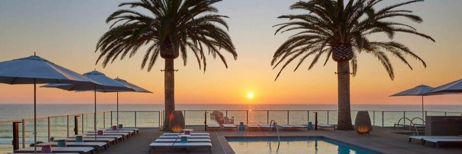 Oceanfront pool at Mission Pacific Beach Resort in Oceanside with lounge chairs, palm trees, umbrellas, and a Pacific sunset.