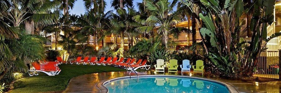 Tropical swimming pool at Ocean Palms Beach Resort in Carlsbad with colorful chairs, red loungers, palm trees, and lush greenery.